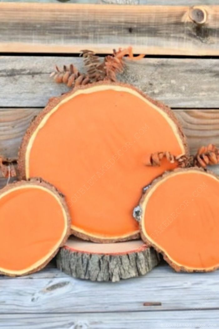 Rustic orange painted wood slices arranged in a pumpkin shape with pinecone accents, set against weathered wooden planks.