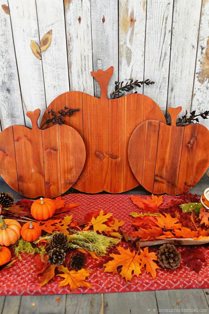 Three wooden pumpkin decorations with autumn leaves and pinecones arranged on weathered wood with a patterned cloth backdrop.