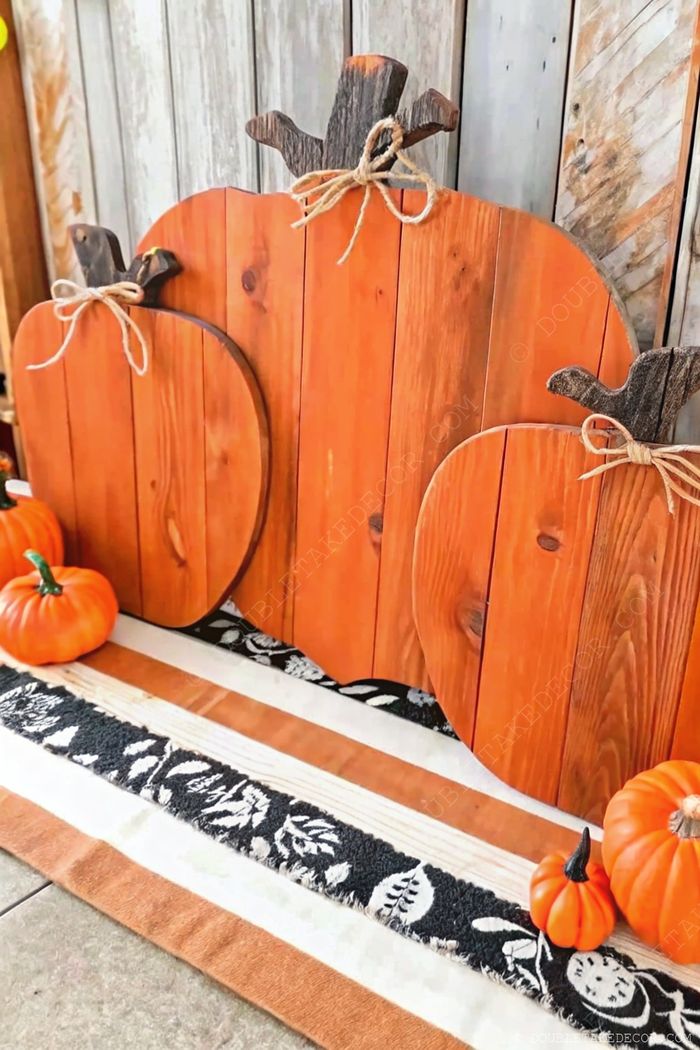 Orange wooden pumpkin decor with twine-wrapped stems displayed alongside mini real pumpkins on an earthy leaf-patterned runner against a rustic wooden backdrop.