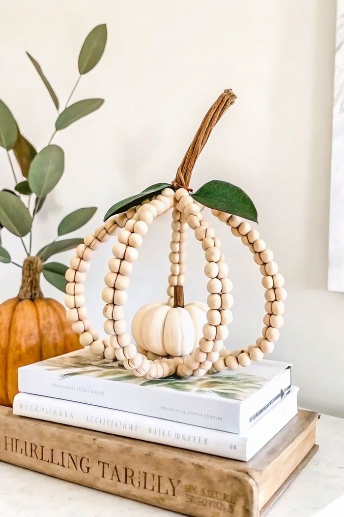 Decorative wooden bead pumpkin with twig stem arranged on stacked books, surrounded by green leaves and a traditional orange pumpkin