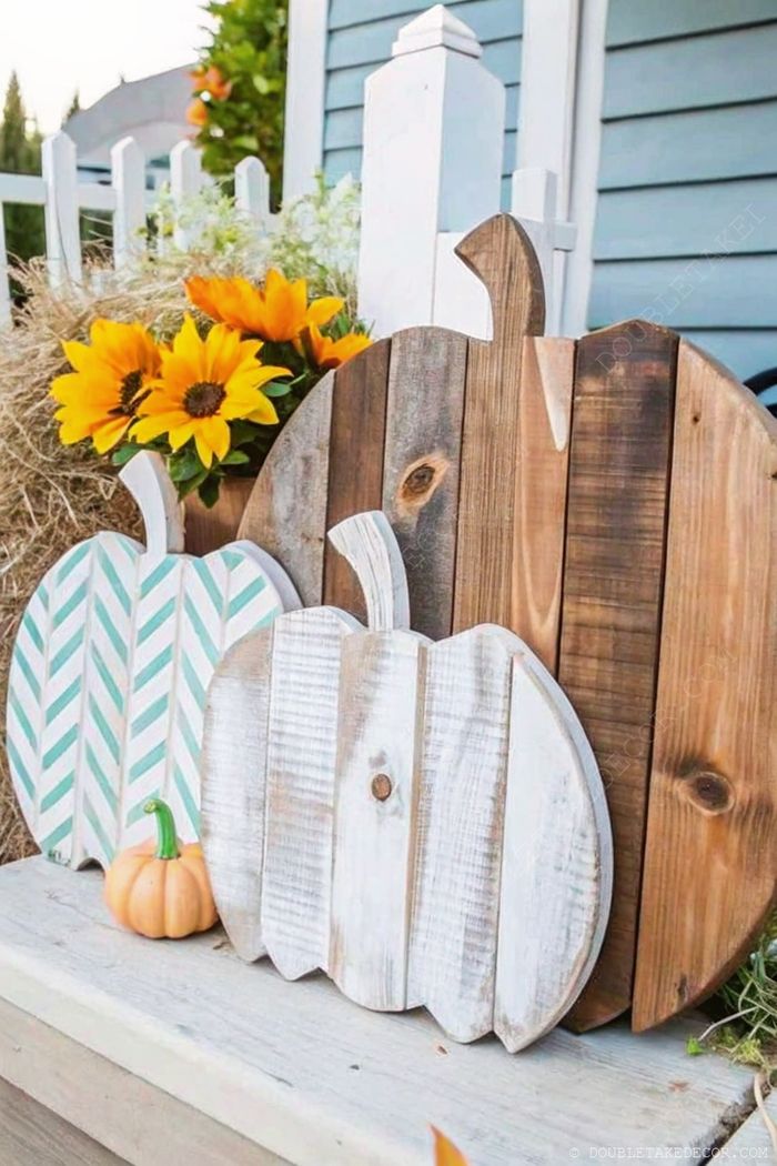 Three decorative wooden pumpkins with varied finishes beside bright sunflowers, set against a blue house and white fence for fall decor.