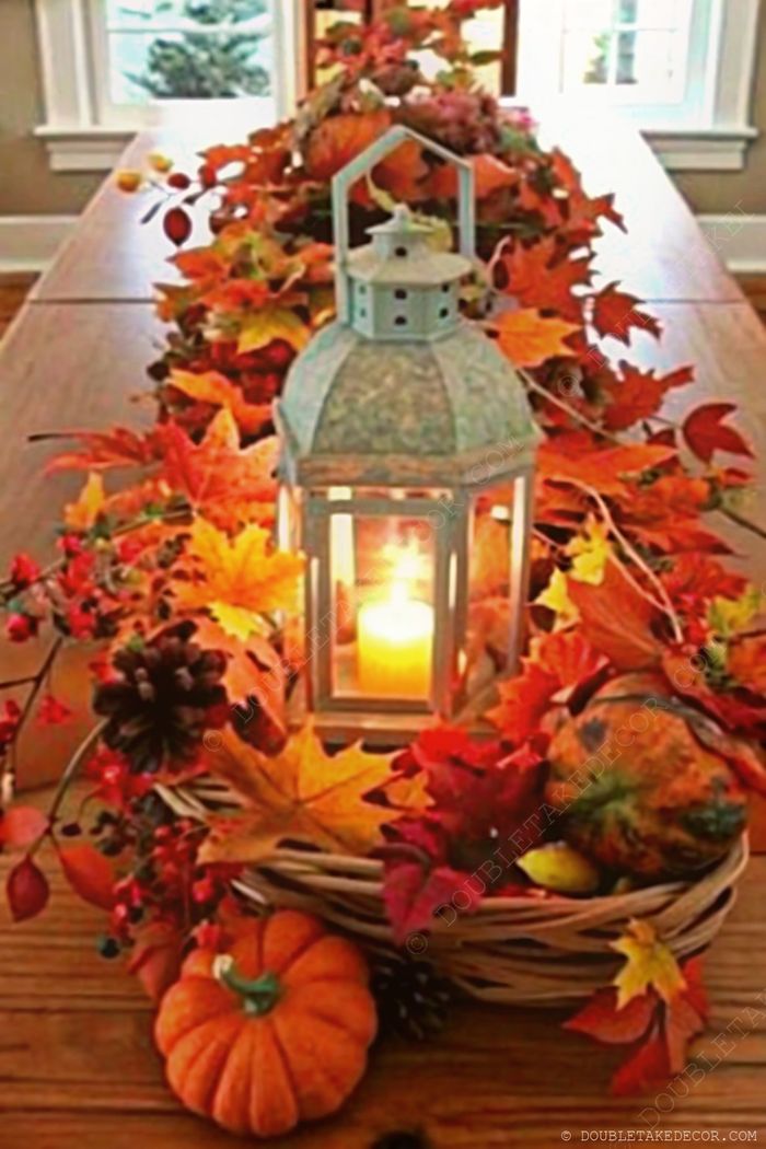 Warm fall centerpiece with glowing lantern, colorful autumn leaves, small pumpkins and pinecones in a woven basket on wooden table.