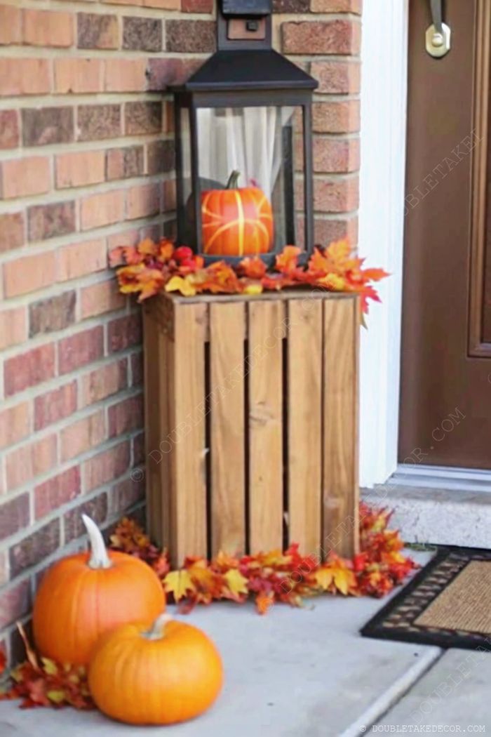 Autumn home entrance with wooden crate, orange pumpkins, colorful leaves, and a decorative lantern on stone steps with a seasonal doormat.