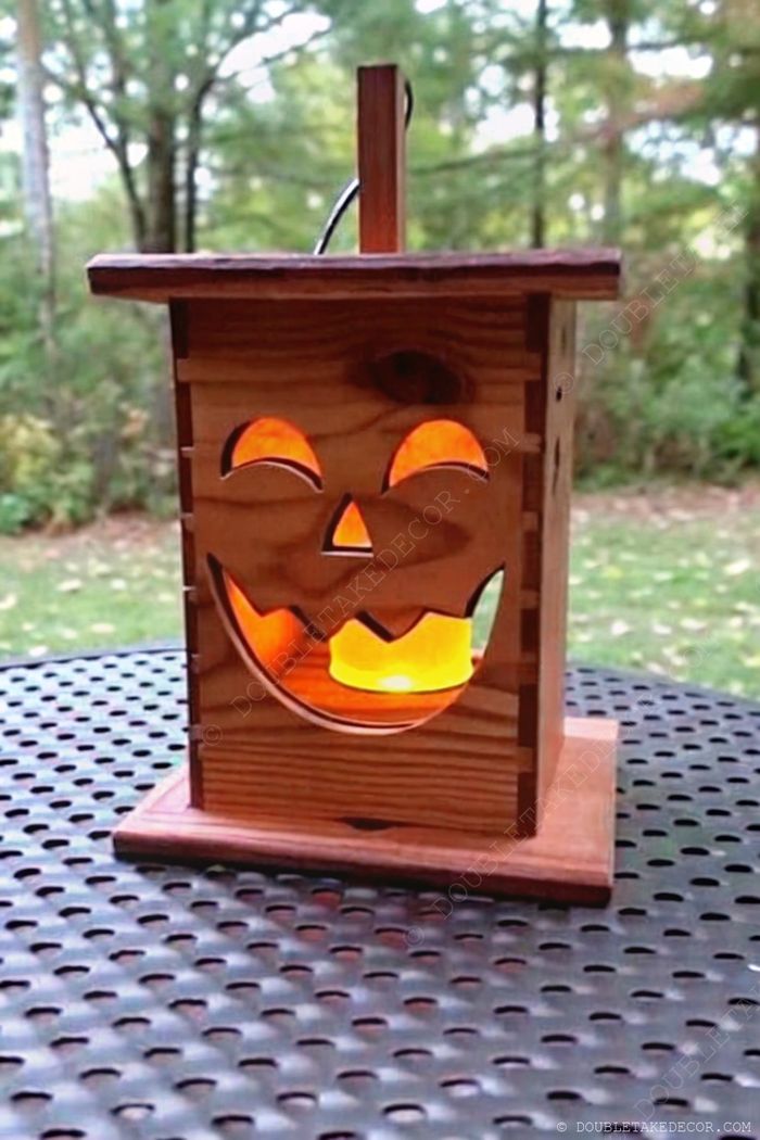 Charming wooden lantern with carved pumpkin face emitting warm light, placed on metal table with green foliage backdrop, perfect for fall decor.