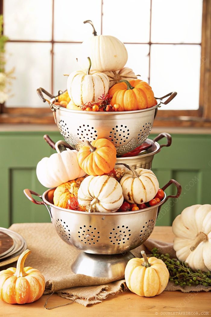 Stacked metal colanders showcasing colorful pumpkins and gourds, creating a rustic autumn centerpiece on burlap tablecloth.