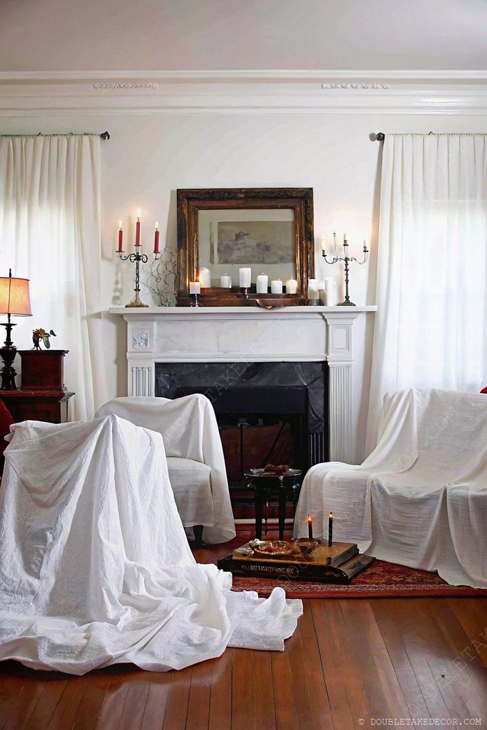 Elegant Halloween living room with white-draped chairs, candlelit mantelpiece, and gold mirror creating a sophisticated spooky ambiance.