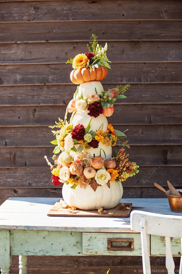 Stacked white pumpkins topped with orange pumpkin, adorned with colorful flowers on rustic teal wooden table, creating elegant autumn decor.
