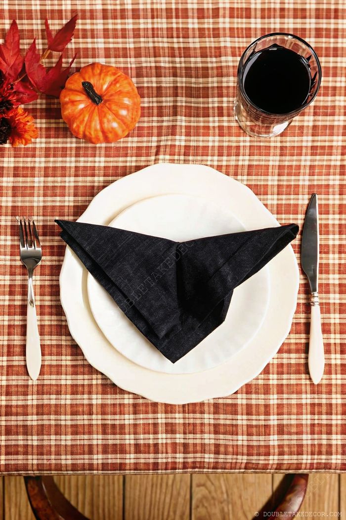 Dining table with orange plaid tablecloth, stacked white plates, black napkin, silverware, and Halloween accents including small pumpkin and fall leaves.