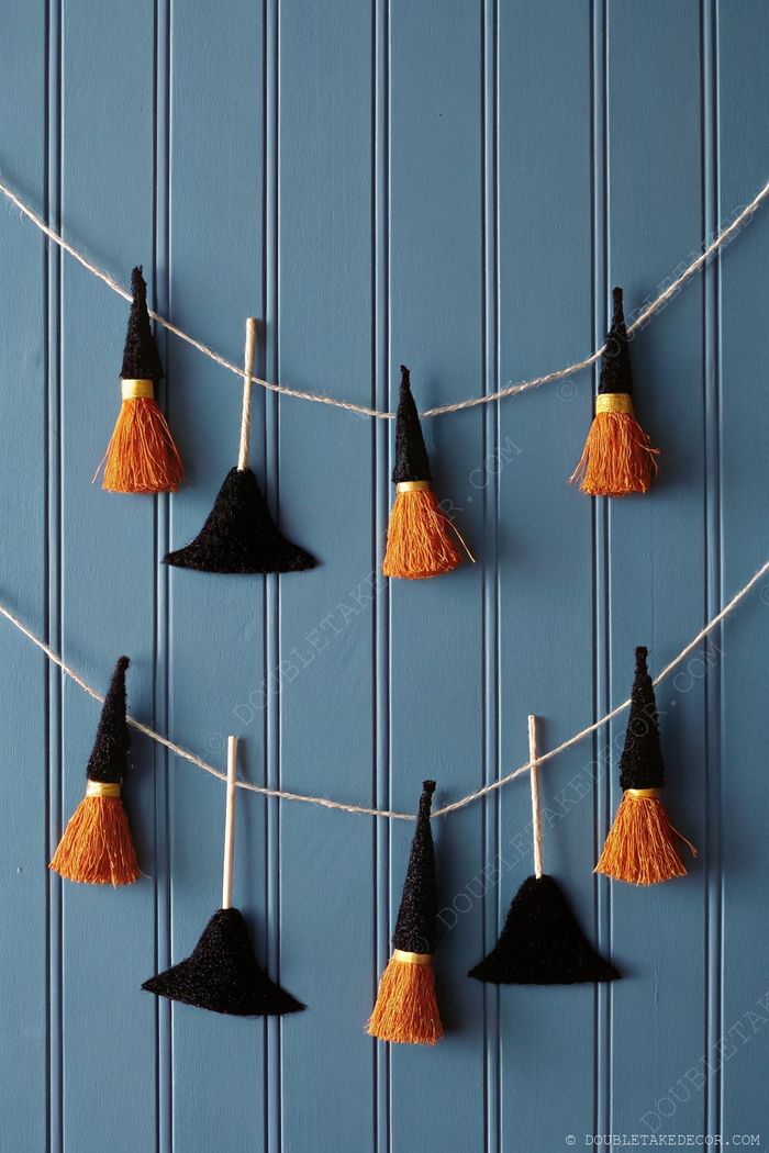 Festive Halloween garland with miniature black witch hats and broomsticks with orange bristles against a blue paneled wall.