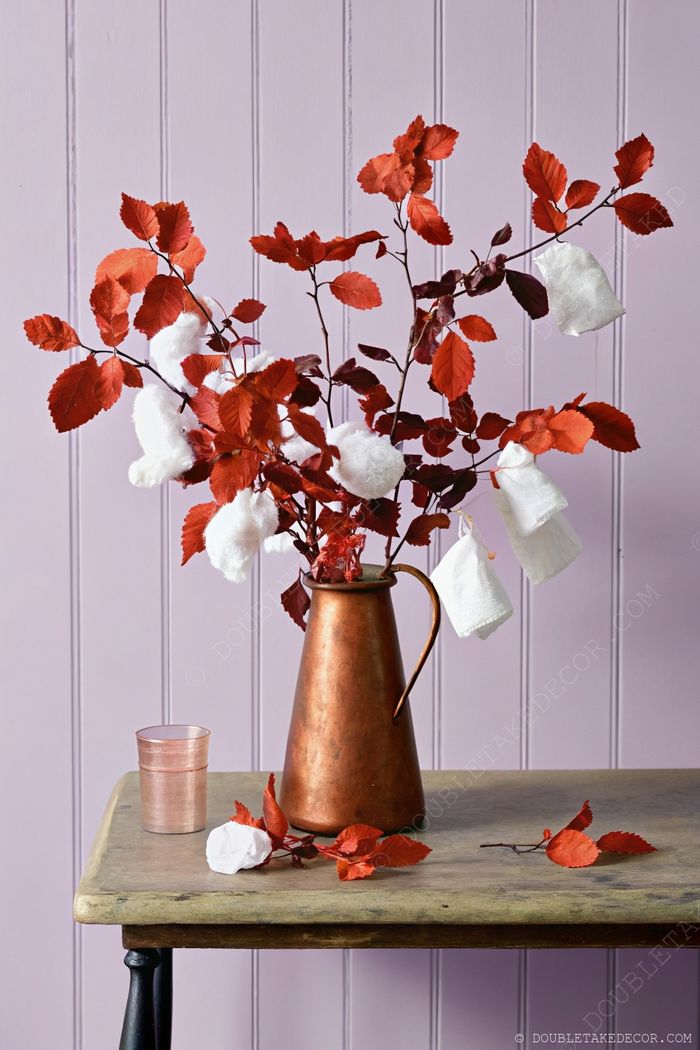 Rustic copper pitcher with vibrant red leaves and white cotton puffs against lilac wall paneling, creating elegant Halloween ambiance