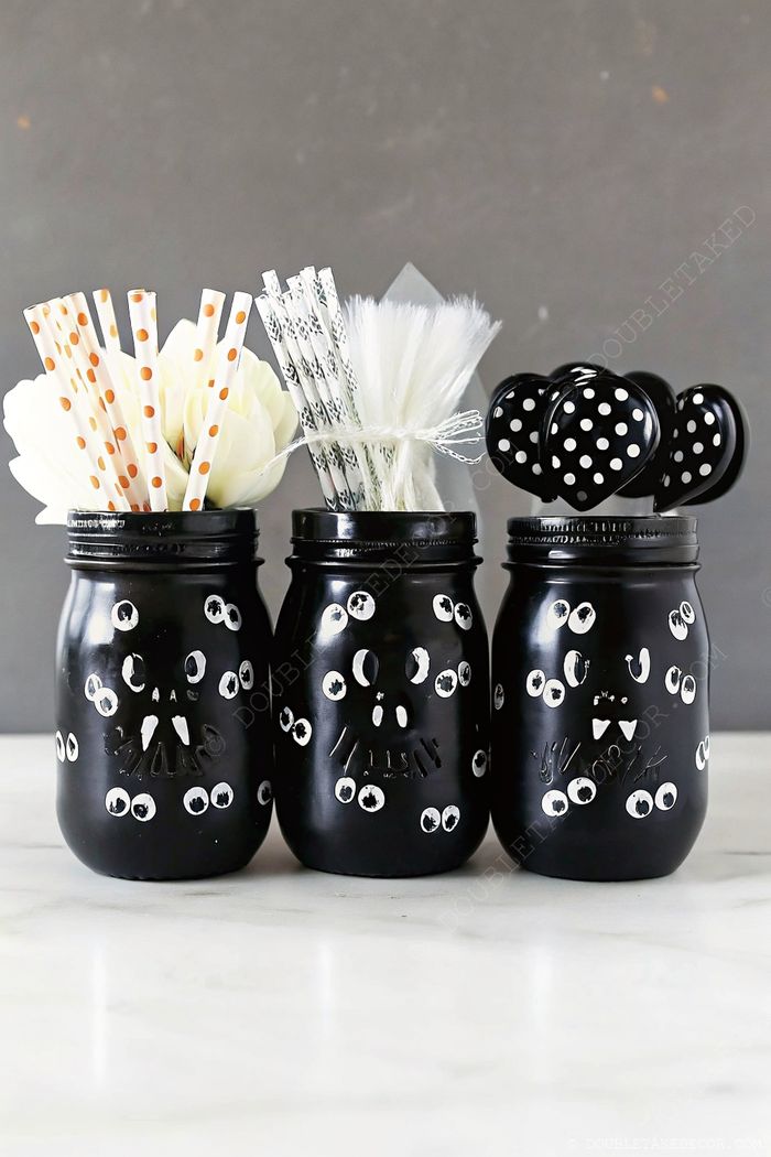 Three black mason jars with white googly eyes, filled with orange striped straws, white decorative materials, and polka-dot items.