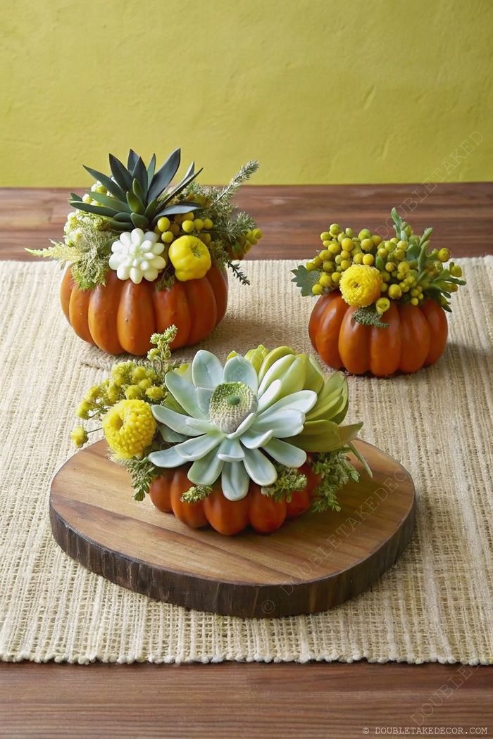 Autumn table centerpiece with three pumpkins decorated with vibrant succulents and yellow flowers on a woven runner with rustic wooden elements.